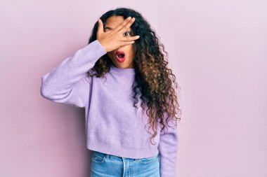 Gündelik giysiler giyen genç Latin kadın şok içinde yüzünü ve gözlerini eliyle kapatıyor, parmaklarından korkuyla bakıyor. 