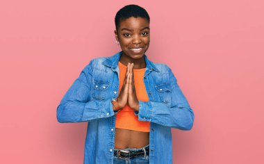 Afro-Amerikalı genç bir kadın günlük kıyafetler giyip ellerinin birliğiyle dua ediyor af diliyor kendinden emin bir şekilde gülümsüyor.. 