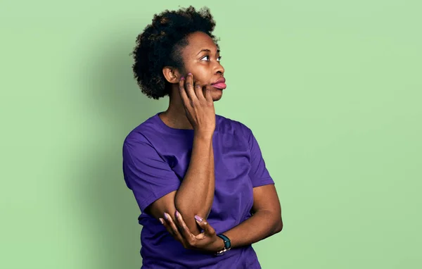 アフリカ系アメリカ人の女性は 質問について考えて顎に手でカジュアルな紫色のTシャツを着てアフロ髪 集中的な表現 思慮深い顔で笑顔 疑わしい概念 — ストック写真