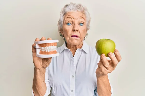 高个子灰发女人 拿着绿色苹果和假牙 毫无头绪 表情混乱 怀疑概念 — 图库照片