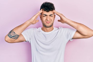 Latin asıllı genç bir adam, günlük beyaz tişört giyiyor, eli başının üstünde, stresten baş ağrısı çekiyor. migren ağrısı. 