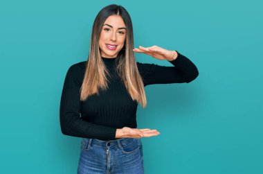 Latin kökenli genç bir kadın, gündelik kıyafetler giyiyor. Elleriyle işaret ediyor. Ölçü sembolü. Kameraya bakıp gülümsüyor. ölçüm kavramı. 