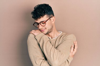 Gündelik kıyafetler ve gözlük takan genç İspanyol adam kendine sarılıyor mutlu ve pozitif, kendinden emin gülümsüyor. Sevgi ve özgüven. 