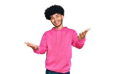 Young african american man with afro hair wearing casual pink sweatshirt smiling cheerful offering hands giving assistance and acceptance.  clipart