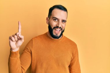 Sakallı genç adam sıradan bir kazak giyiyor. Bir numaralı parmağını gösterip gülümsüyor. Kendinden emin ve mutlu.. 