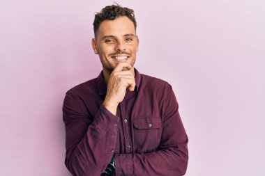 Gündelik giysiler giyen genç İspanyol adam kameraya kollarını kavuşturup çenesine dokunarak gülümsüyor. Olumlu düşünmek. 