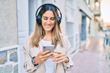 Beyaz kadın şehirde akıllı telefon ve kulaklık kullanarak mutlu bir şekilde gülümsüyor..