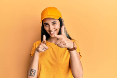 Teslimat üniforması giyen ve şapkalı genç İspanyol kadın mutlu ve komik yüzüyle kamerayı işaret ediyor. iyi enerji ve titreşimler. 