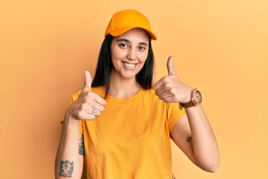Teslimat üniforması giyen ve şapka takan genç İspanyol kadın elleriyle olumlu jest yapıyor, gülümsüyor ve mutlu. Neşeli ifade ve kazanan jest. 