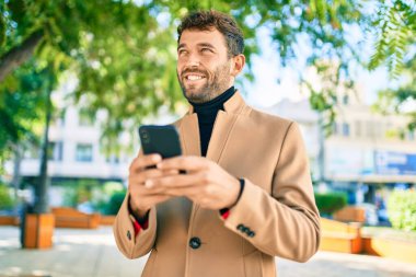 Zarif bir ceket giyen yakışıklı bir iş adamı akıllı telefon kullanarak dışarıda mutlu bir şekilde gülümsüyor.