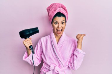 Duşta bornoz giyen, kurutma makinesi kullanan genç İspanyol kadın parmak ucunda ağzı açık gülümsüyor. 