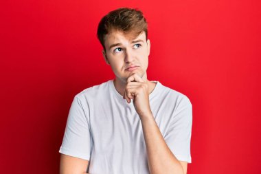 Beyaz günlük tişört giyen genç beyaz bir adam şüpheye odaklanmış, çenesinde parmak olan ve kafasını kaldırıp, 