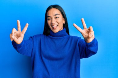 Kış süveteri giymiş genç Asyalı bir kadın her iki elinin de parmaklarını göstererek gülümseyerek zafer işareti yapıyor. İki numara.. 