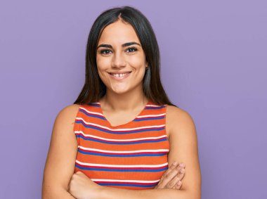 Genç, esmer, sıradan kıyafetler giyen mutlu yüzlü, kollarını kavuşturup kameraya bakan bir kadın. pozitif kişi. 