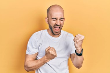 Gündelik beyaz t-shirt giyen genç kel adam şaşkınlık ve şaşkınlık içinde kolları havada ve gözleri kapalı bir şekilde başarıyı kutluyor. Kazanan konsept. 