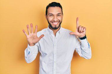 Sakallı İspanyol bir adam iş gömleği giyiyor ve yedi numaralı parmaklarıyla işaret ediyor. Kendinden emin ve mutlu bir şekilde gülümsüyor.. 