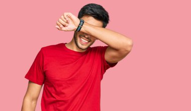 Genç yakışıklı adam, sıradan kırmızı tişörtler giyiyor. Gözleri kollarıyla kaplıyor. Neşeli ve komik gülümsüyor. kör kavram. 