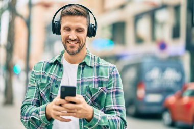 Genç beyaz adam şehirde akıllı telefon ve kulaklık kullanarak mutlu bir şekilde gülümsüyor..