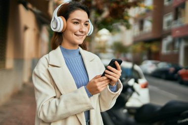 Genç İspanyol kadın şehirde akıllı telefon ve kulaklık kullanarak mutlu bir şekilde gülümsüyor..