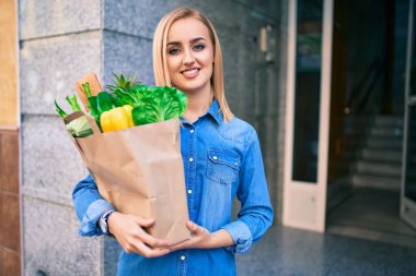 Genç sarışın kız gülümsüyor. Elinde alışveriş poşeti, şehirde duruyor..
