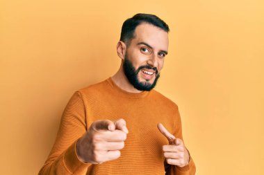 Kış süveteri giyen sakallı genç adam mutlu ve komik yüzüyle kamerayı işaret ediyor. iyi enerji ve titreşimler. 