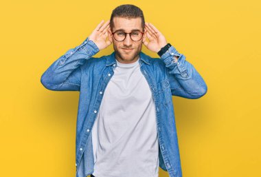 Günlük kıyafetler giyen genç beyaz bir adam iki elini de kulak hareketlerini duymaya çalışıyor. Dedikoduya meraklı. İşitme sorunu, sağır 