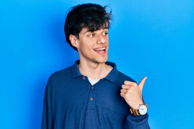 Yakışıklı hippi genç adam mavi arka planda mahsur kalmış mutlu bir yüzle gülümsüyor ve başparmağıyla yan tarafı işaret ediyor.. 