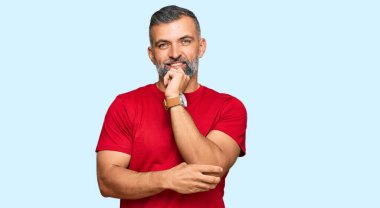 Orta yaşlı, sıradan kıyafetler giyen, kameraya kendinden emin, kolları çapraz ve çenesi kalkık bir şekilde bakan yakışıklı bir adam. Olumlu düşünmek. 