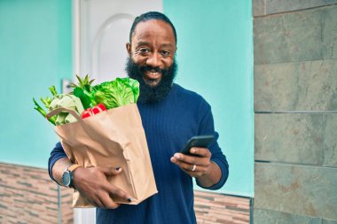 Sakallı Afrikalı Amerikalı adam elinde süpermarketten alınmış kağıt torbalarla akıllı telefon kullanıyor.