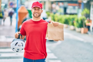 Beyaz bir teslimatçı, elinde kağıt torbayla şehirde mutlu bir şekilde gülümsüyor..