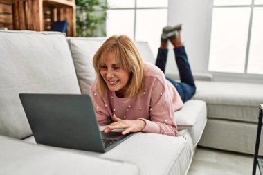 Orta yaşlı sarışın kadın evdeki kanepede dizüstü bilgisayar kullanıyor..