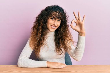 Latin asıllı genç kız gündelik giysiler içinde masada oturmuş gülümsüyor ve el ve parmaklarıyla 