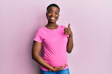 Hamile karnına dokunarak mutlu bir jest yapan, bebek bekleyen Afrikalı Amerikalı genç bir kadın. Kameraya bakıp başarıyı gösteren ifade. 