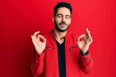 Kırmızı deri ceket giyen, gözleri kapalı gülümseyen genç İspanyol adam parmaklarıyla meditasyon hareketi yapıyor. yoga konsepti. 