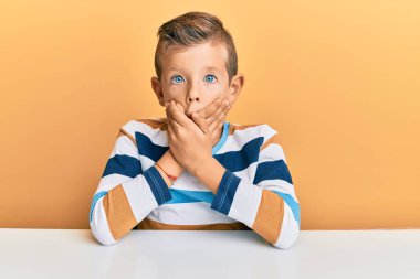 Masada oturan beyaz giysili sevimli çocuk ağzı yanlışlıkla ellerle kapatırken şok oldu. gizli kavram. 