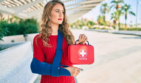Young blonde girl with serious expression injuried with arm sling and holding first aid kit at the park.