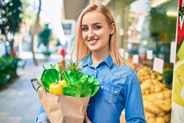 年轻的金发姑娘笑着高高兴兴地拿着食品杂货纸袋站在超市前 — 图库照片