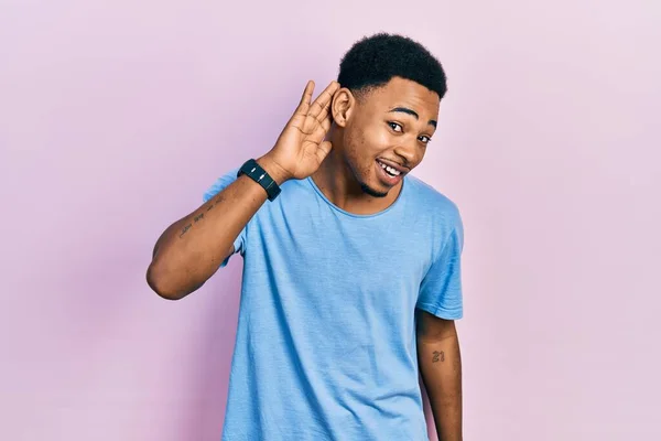 噂やゴシップに耳を傾ける耳の上に手で笑みを浮かべてカジュアルな青のTシャツを着て若いアフリカ系アメリカ人の男性 聴覚障害の概念 — ストック写真