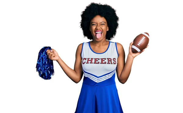 身穿啦啦队队长制服的年轻的非洲裔美国女人 手里拿着花环和足球 带着滑稽的表情伸出舌头 高兴极了 — 图库照片