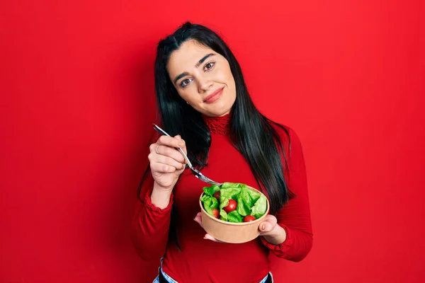 真剣な表情でリラックスしたサラダを食べている若いヒスパニック系の女の子 カメラを見ているだけで — ストック写真