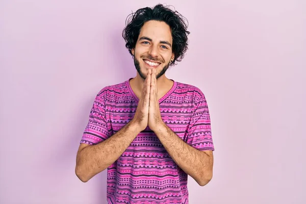 手で一緒に祈るカジュアルなピンクのTシャツを身に着けているハンサムなヒスパニック系の男は自信を笑顔許しを求めて — ストック写真