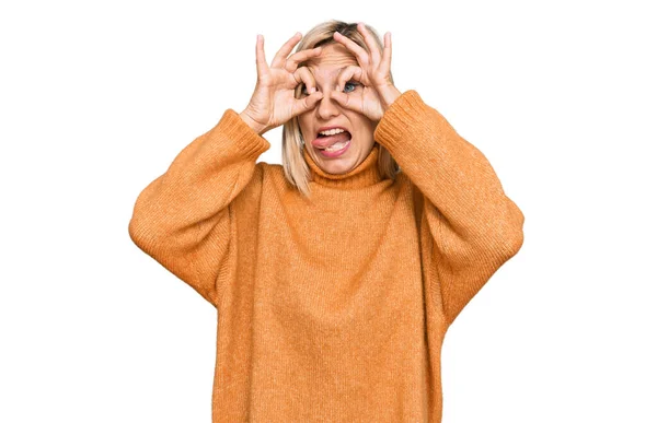 中年的高加索女人穿着宽松的冬季毛衣 像双筒望远镜一样做手势 伸出舌头 眼睛透过手指看过去 疯狂的表达 — 图库照片