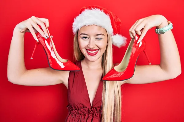 年轻的金发女郎头戴圣诞帽 头戴高跟鞋 眨着眼睛 带着性感的表情 开朗而快乐地看着摄像机 — 图库照片