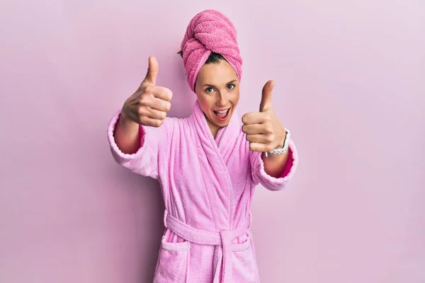 Young blonde woman wearing shower towel cap and bathrobe approving doing positive gesture with hand, thumbs up smiling and happy for success. winner gesture.