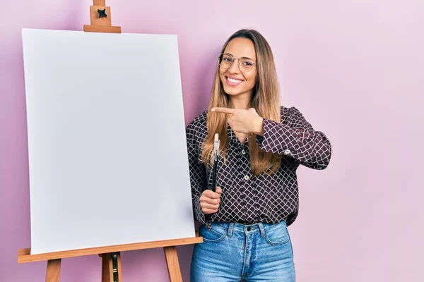 画家のそばに立つ美しいヒスパニック系の女性イーゼルスタンド手と指で幸せなポインティング笑顔ブラシを保持 — ストック写真