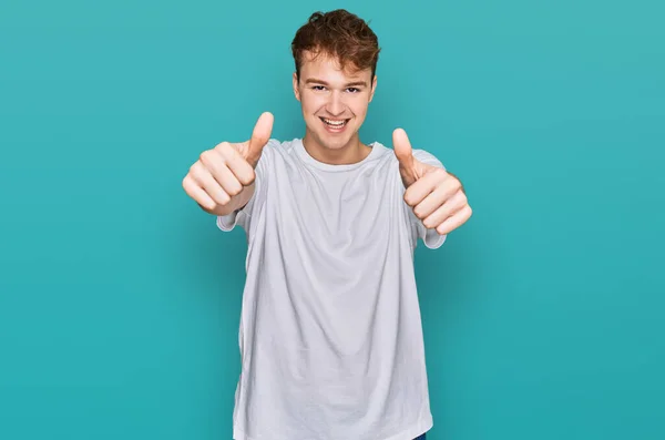 年轻的高加索男人穿着宽松的白色T恤 赞成用手做积极的手势 微笑着竖起大拇指 为成功感到高兴 优胜手势 — 图库照片