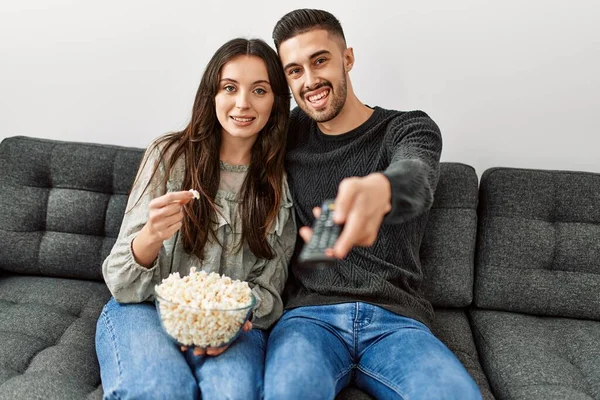 Young hispanic couple watching film and eating popcorn sitting on the sofa at home.