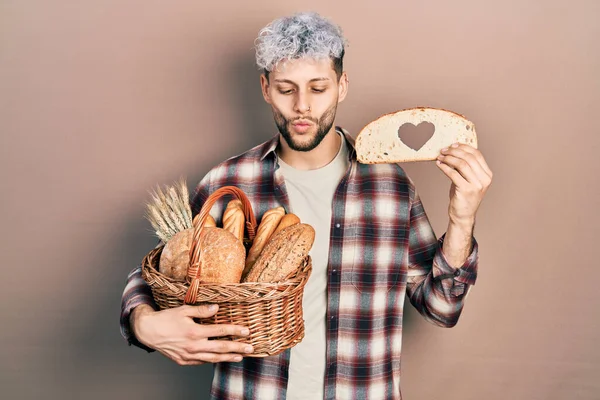 Young hispanic man with modern dyed hair holding wicker basket with bread and loaf with heart symbol making fish face with mouth and squinting eyes, crazy and comical.