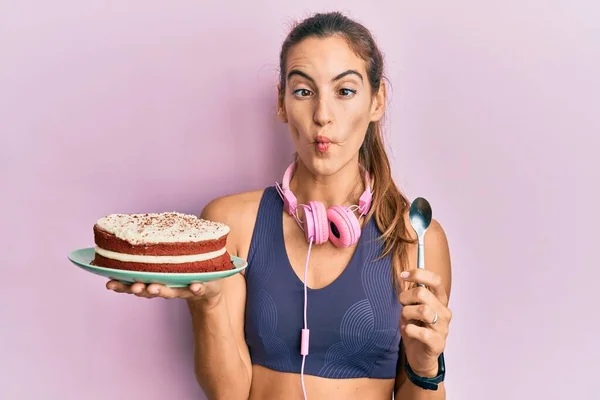 Young beautiful woman wearing gym clothes holding carrot cake making fish face with mouth and squinting eyes, crazy and comical.