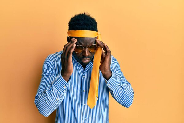 Handsome black man drunk wearing tie over head and sunglasses with hand on head for pain in head because stress. suffering migraine.